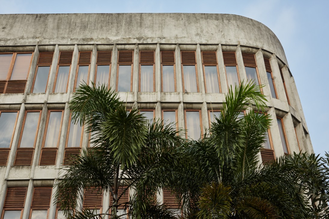 Curved concrete facade of Anjung Salihara building with rows of windows and wooden shutters, palm trees in foreground
