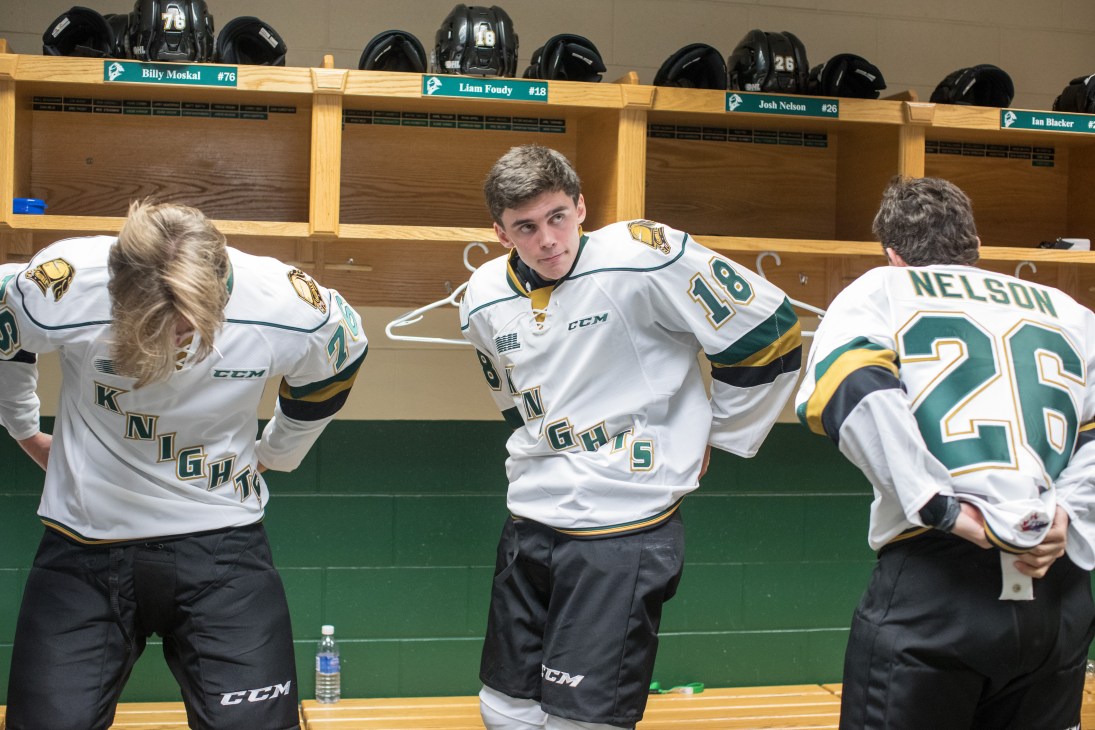 Hockey players in white Knights jerseys getting dressed in wooden locker room stalls
