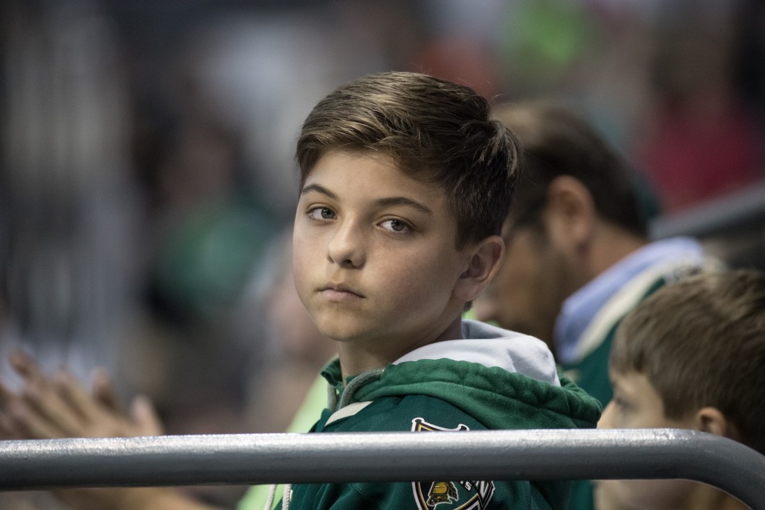 Young boy in green and white hockey jersey sits in arena bleachers looking pensively into the distance