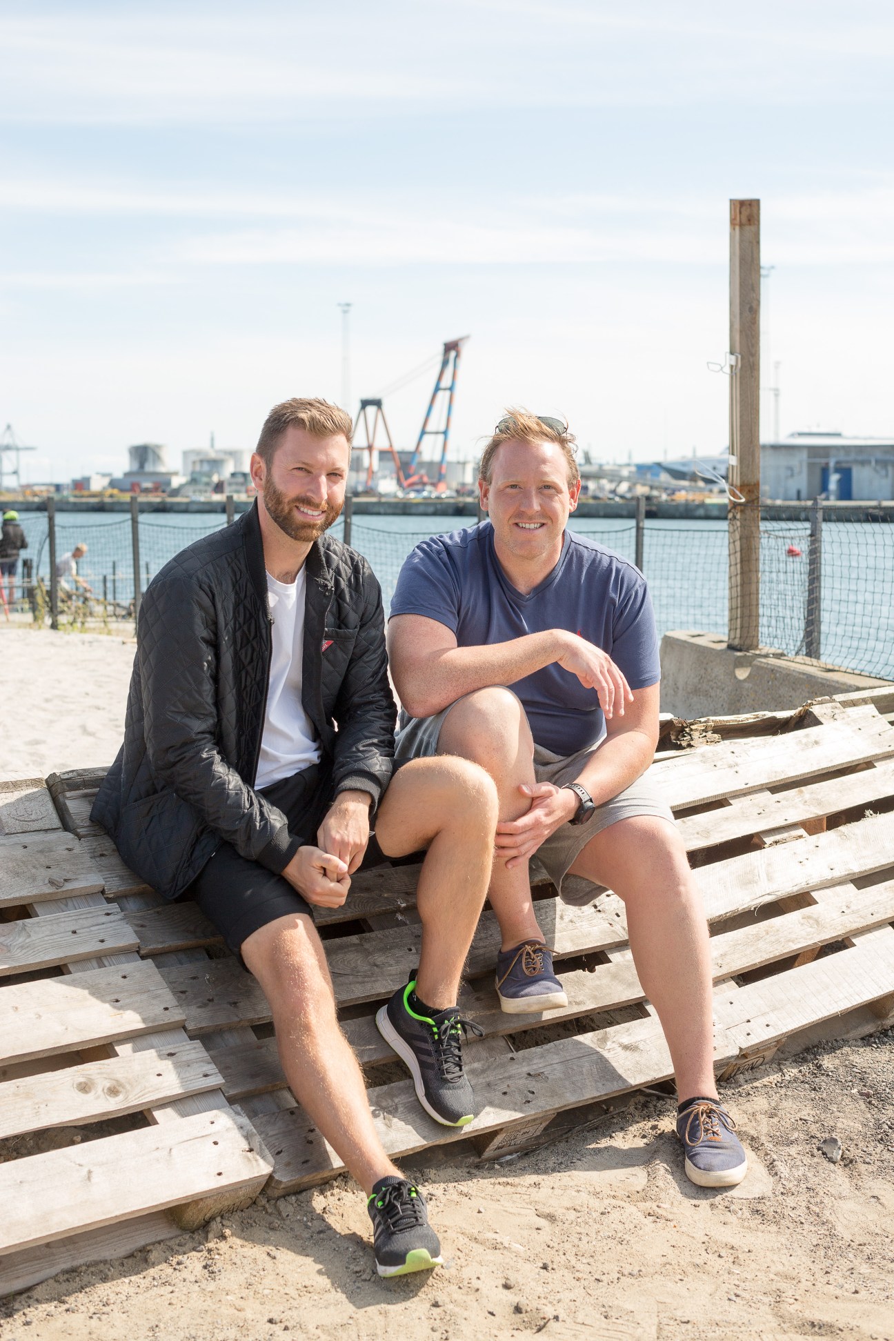 Two men sit on wooden pallets on a beach with a shipping port visible behind them.