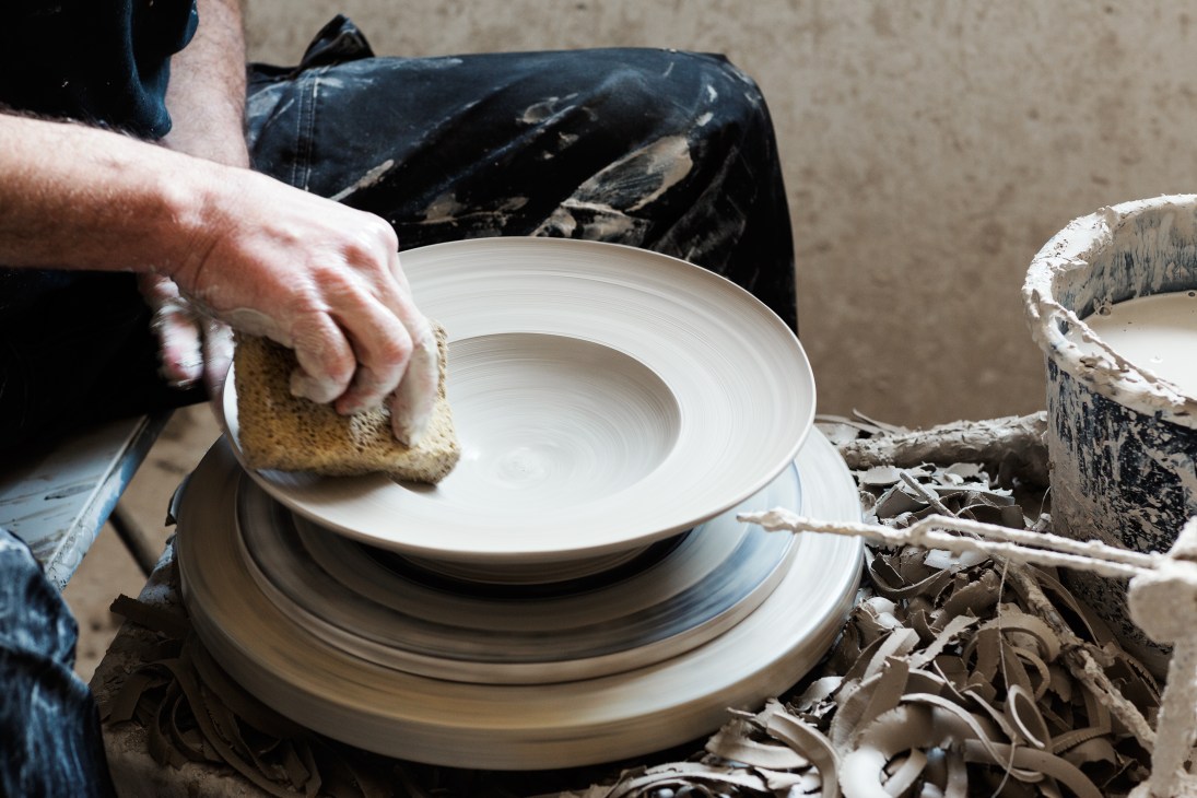 A craftsperson uses a sponge to moisten and smooth a white ceramic dish during the finishing process.