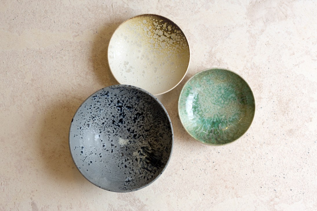 Three handmade ceramic dishes in varying sizes and colors: dark speckled gray, cream with yellow speckles, and green with organic patterns.