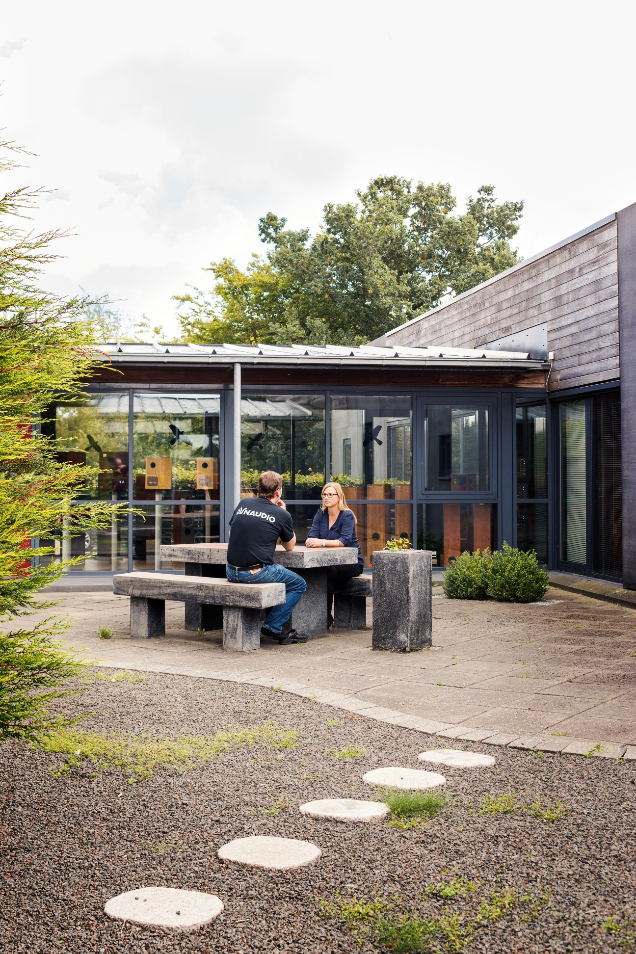Two people sit at a concrete table in a courtyard outside a modern dark-clad building with large windows.