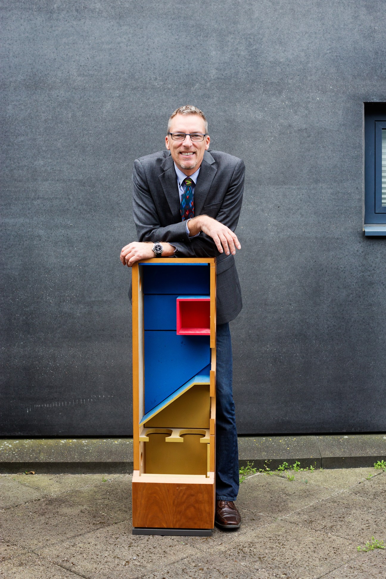 Lars Prisak, Dynaudio's CEO, poses with a colorful wooden loudspeaker model against a gray building facade.