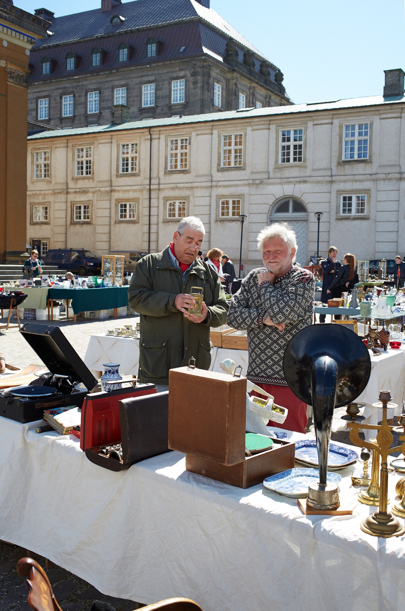 Two men examining antiques at an outdoor market in a European city square.