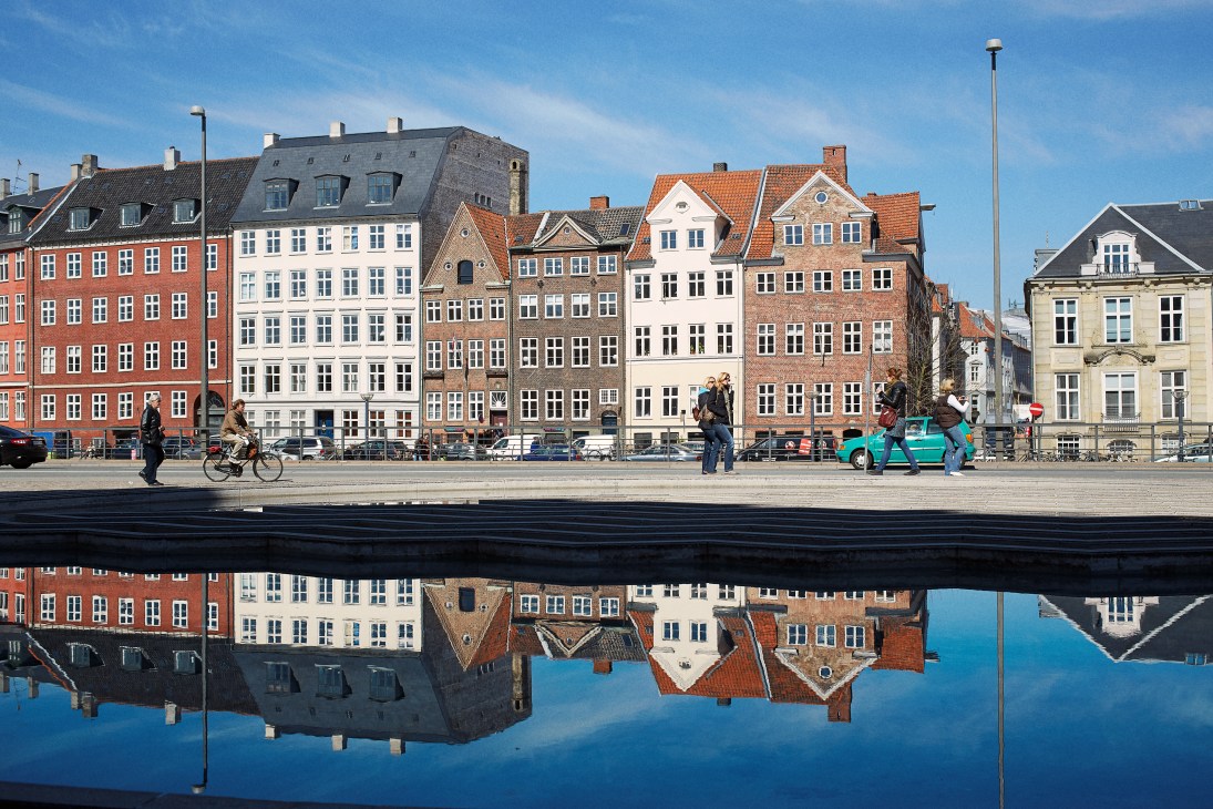 Colorful historic buildings reflected in still water with pedestrians and cyclist on waterfront promenade in Copenhagen.