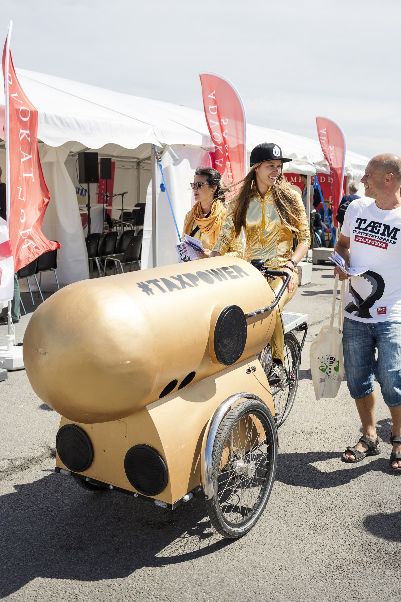 Two women in golden jackets stand with a large golden capsule-shaped vehicle marked with #TAXPOWER at Folkemødet festival.