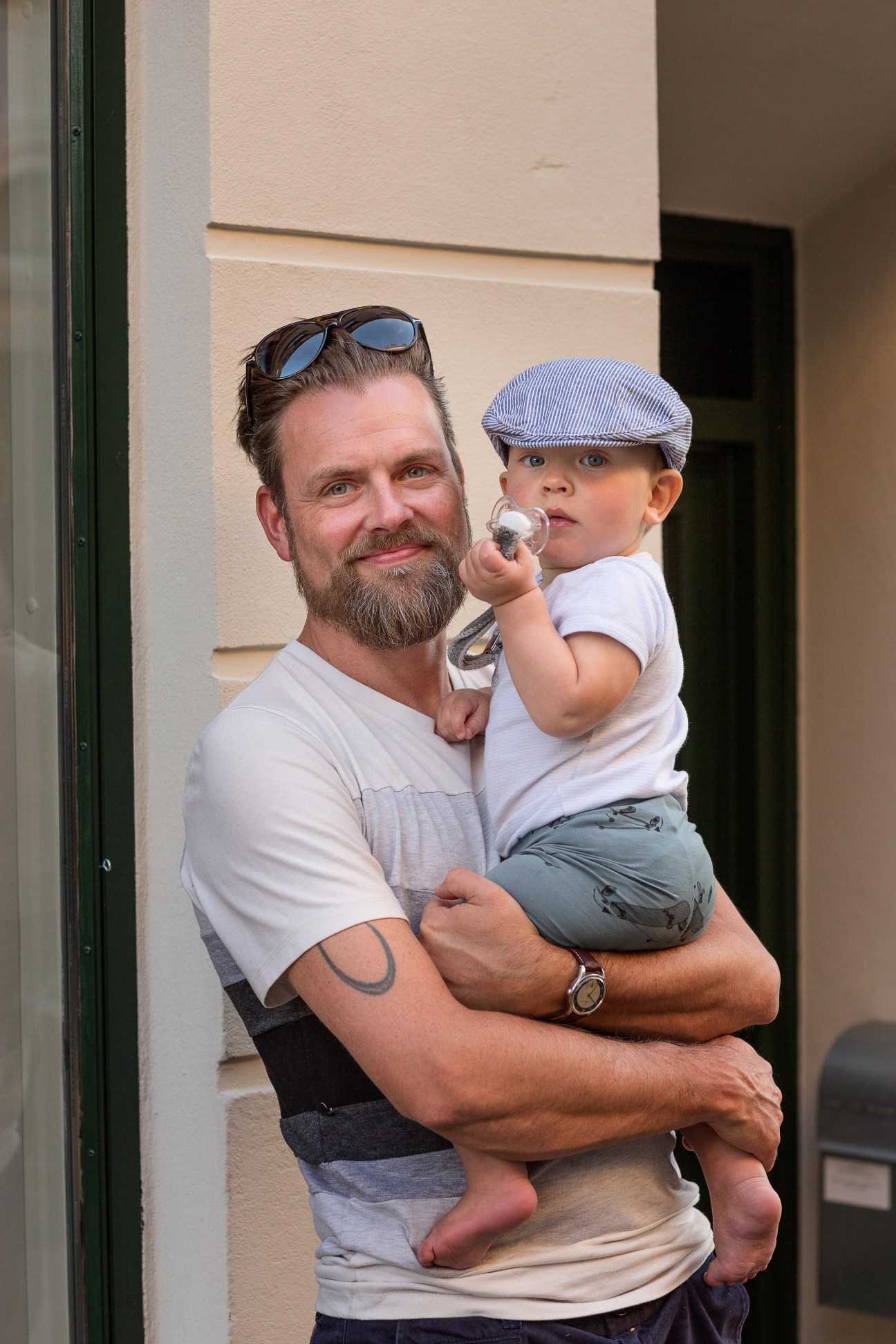A bearded man holding a young child in a blue cap against a cream-colored building wall.