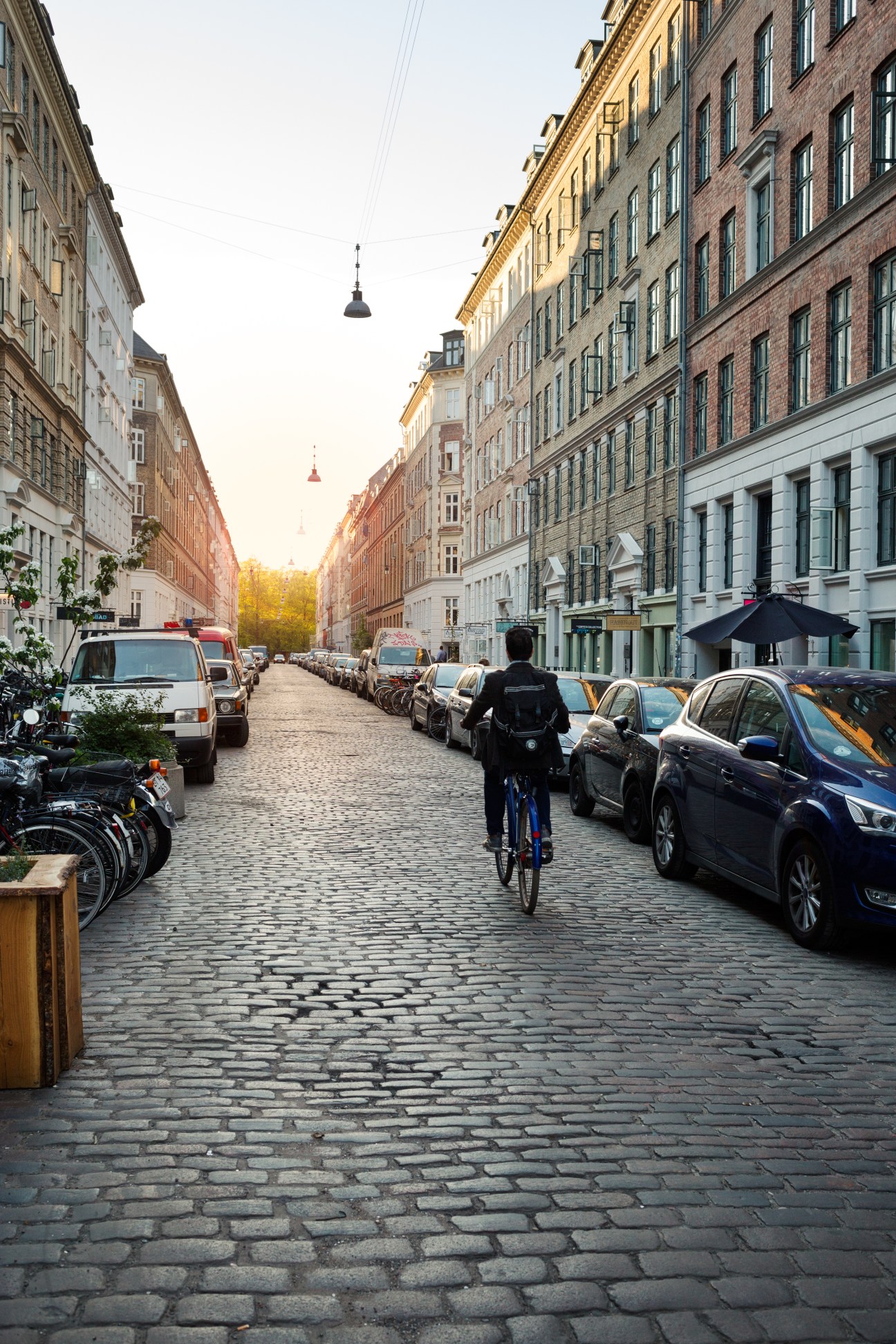 A cyclist rides down a cobblestone street at sunset with parked cars and historic buildings lining both sides.