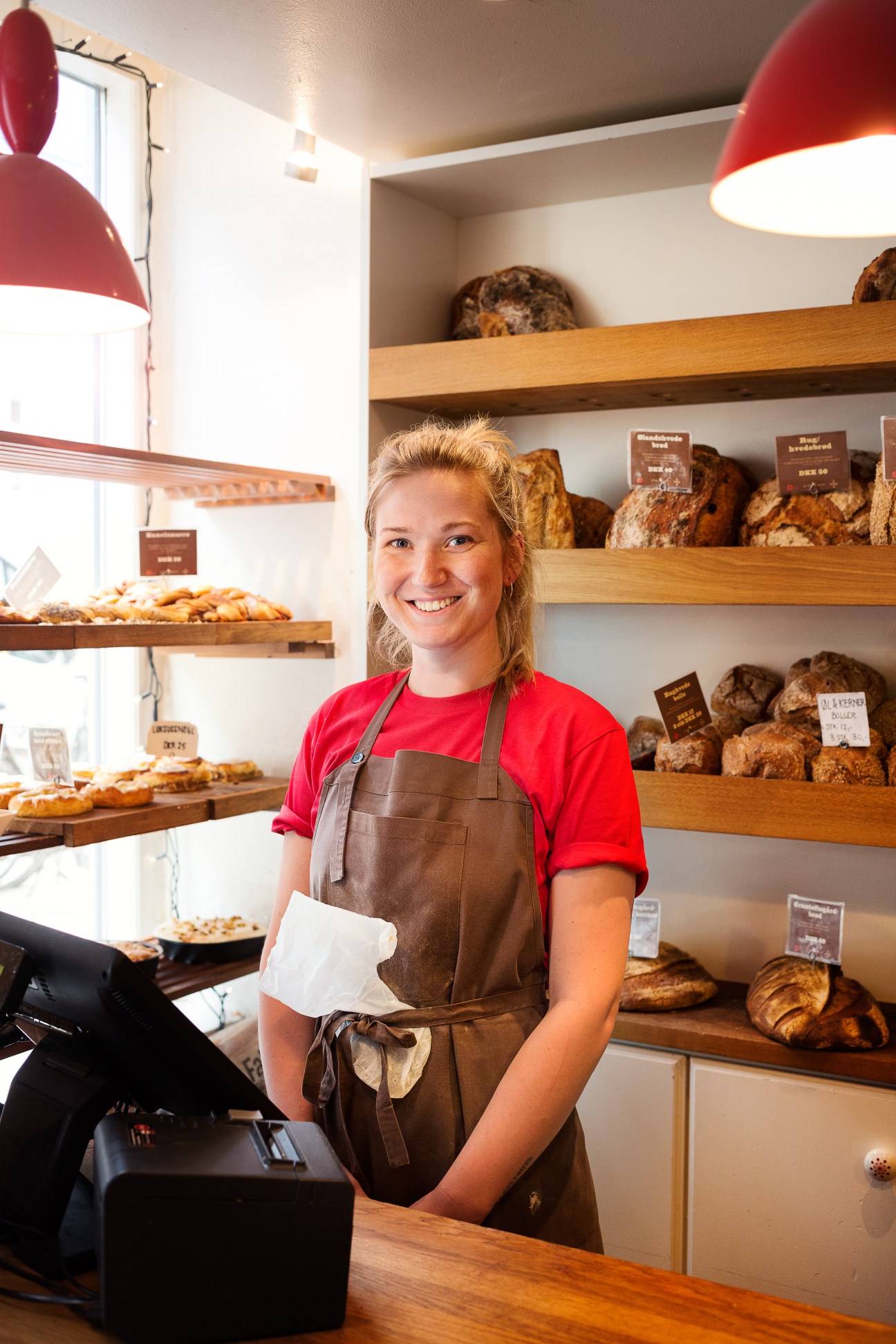 Smiling bakery staff member in brown apron standing in front of shelves displaying fresh breads and pastries.