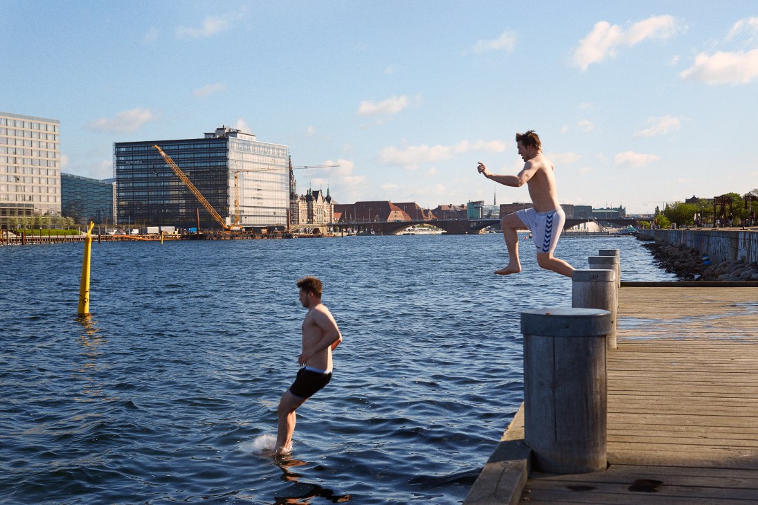 Young people swimming and jumping from a wooden dock into Copenhagen's harbor with modern buildings in the background.