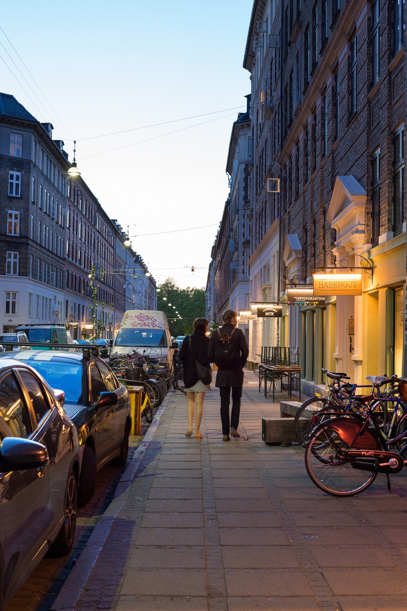 Jaegersborggade street at dusk with illuminated storefronts, parked bicycles, and two pedestrians walking.