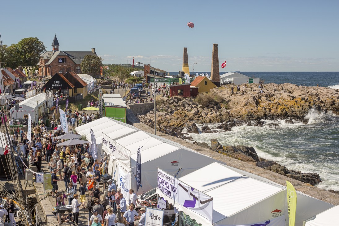 Aerial view of Allinge harbour with festival tents, crowds, rocky coastline and inflatable pink pig in sky.