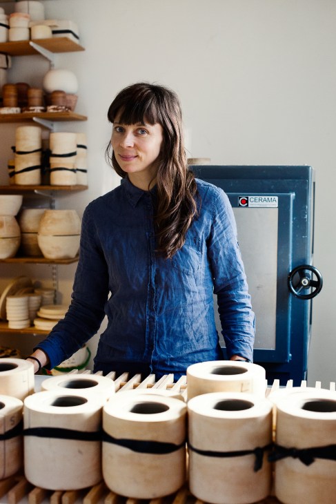 Potter Mette Duedahl stands in her studio surrounded by cream-colored ceramic vessels and shelves of pottery.