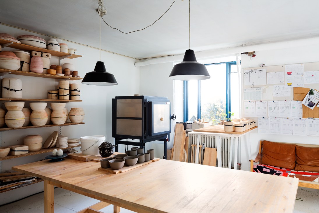 A bright pottery studio with wooden shelves displaying cream and earth-toned ceramic vessels, a kiln, work table, and planning boards on the wall.