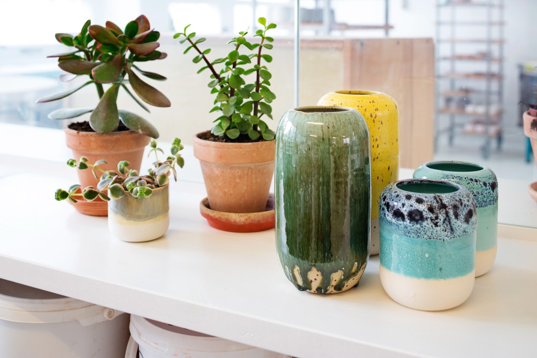 Colorful ceramic vessels and planters with succulents displayed on a white shelf by a window.