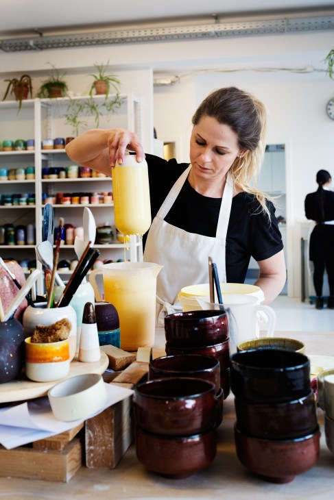 Potter pouring yellow glaze into ceramic vessels in a bright studio workspace.