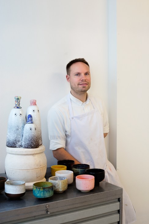 Anders Arhoj stands next to a display table showcasing his ceramic creations, including white ghost figurines and colorful pottery bowls.