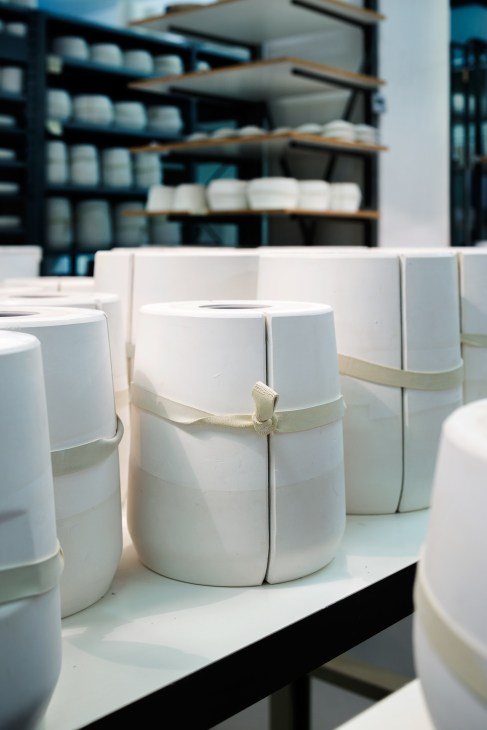 White porcelain vessels wrapped with cream ribbons displayed on a table with shelving in background.