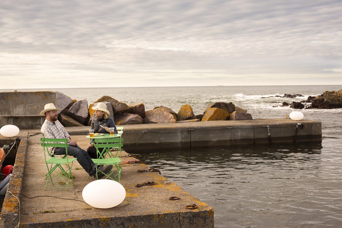 Two people sit on green camping chairs on a harbour wall by the sea, with white pendant lights and food.