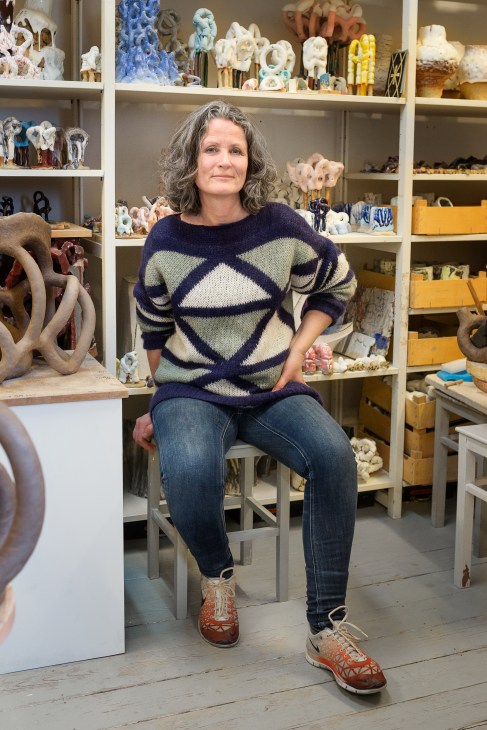 Woman sitting in a ceramics studio with shelves of pottery pieces behind her.