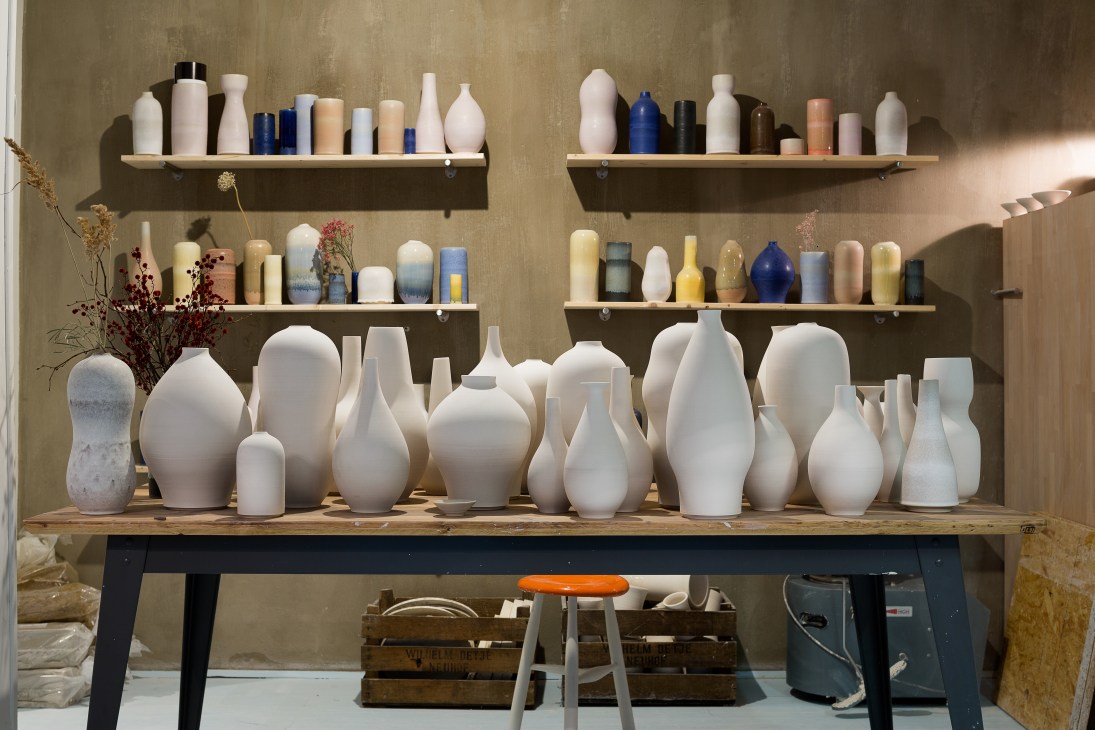 White ceramic vessels arranged on a wooden table with shelves of colorful pottery bottles displayed on the wall behind them at Tortus Copenhagen.
