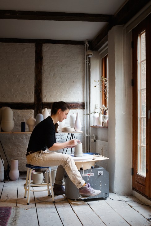 An apprentice sits at a pottery wheel working on a ceramic vessel in a bright studio space with white vessels displayed on shelves behind.