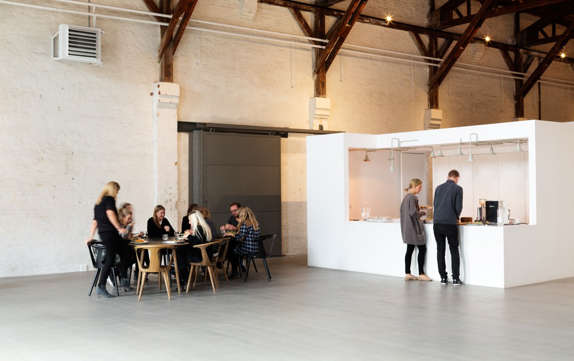 &Tradition staff gather around a dining table in the showroom's kitchen area with white cube structures and exposed wooden beams.
