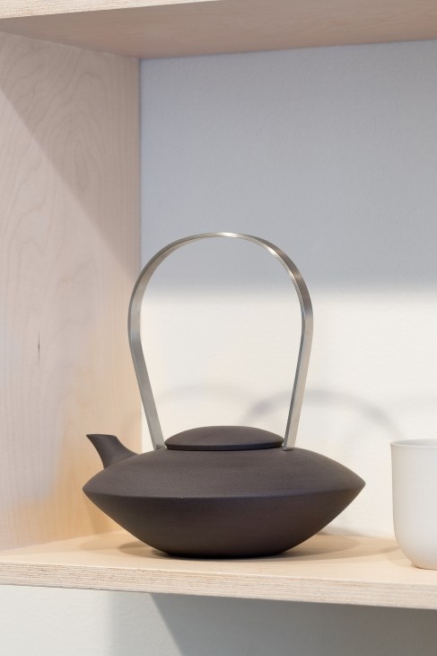 A black porcelain teapot with a metal handle and minimalist design displayed on a wooden shelf.