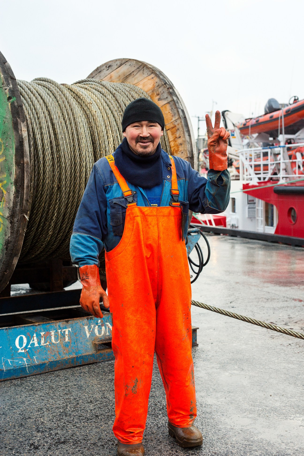A Nuuk fisherman in orange waders and blue jacket stands at a harbor with fishing vessels behind him.