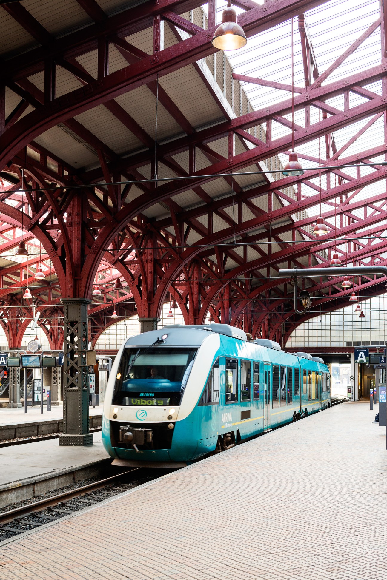 Turquoise and white train at Aarhus Central Station platform beneath red iron beams and glass roof.