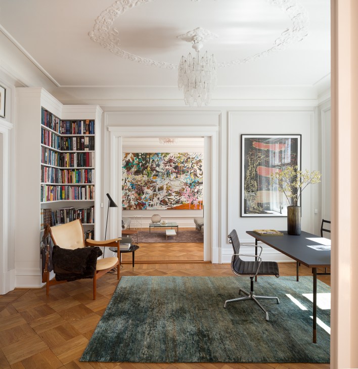 Bright apartment interior with bookshelves, desk, artwork, and modern furnishings across connected rooms.