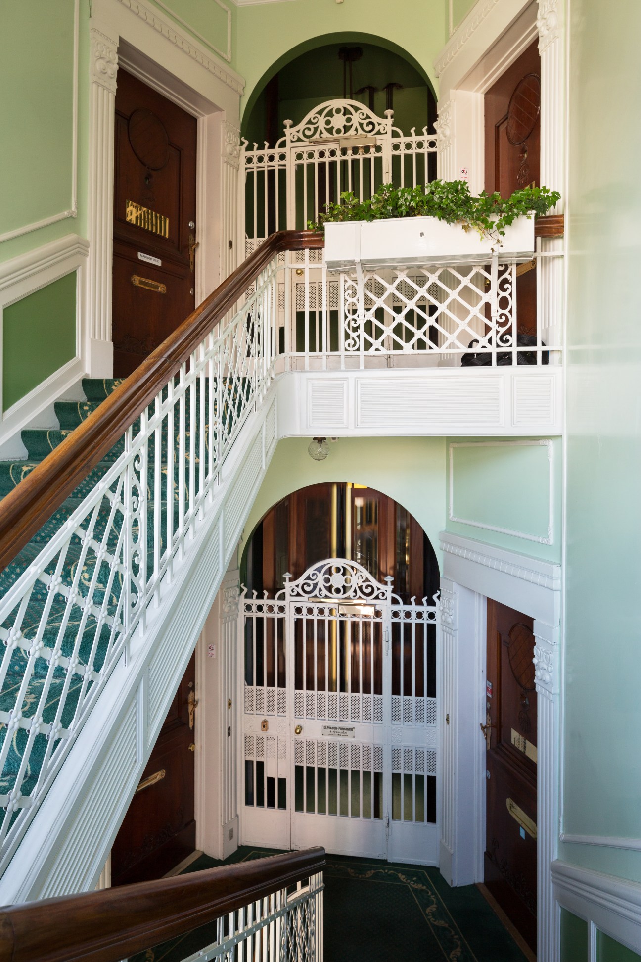 Elegant multi-story stairwell with white railings, green walls, dark wooden doors, and ornate arched doorways in a historic Copenhagen building.