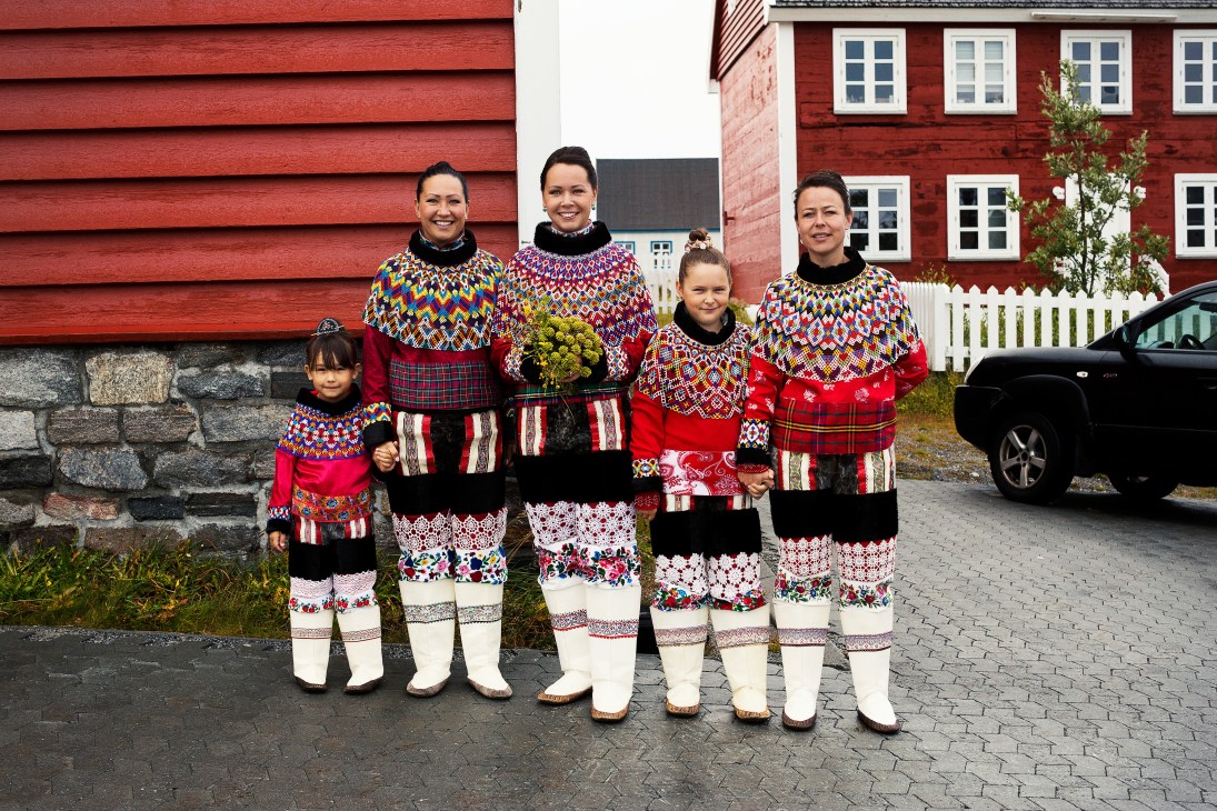 A wedding party of five people wearing colorful traditional Greenlandic costumes with patterned sweaters and boots standing in front of red colonial buildings.