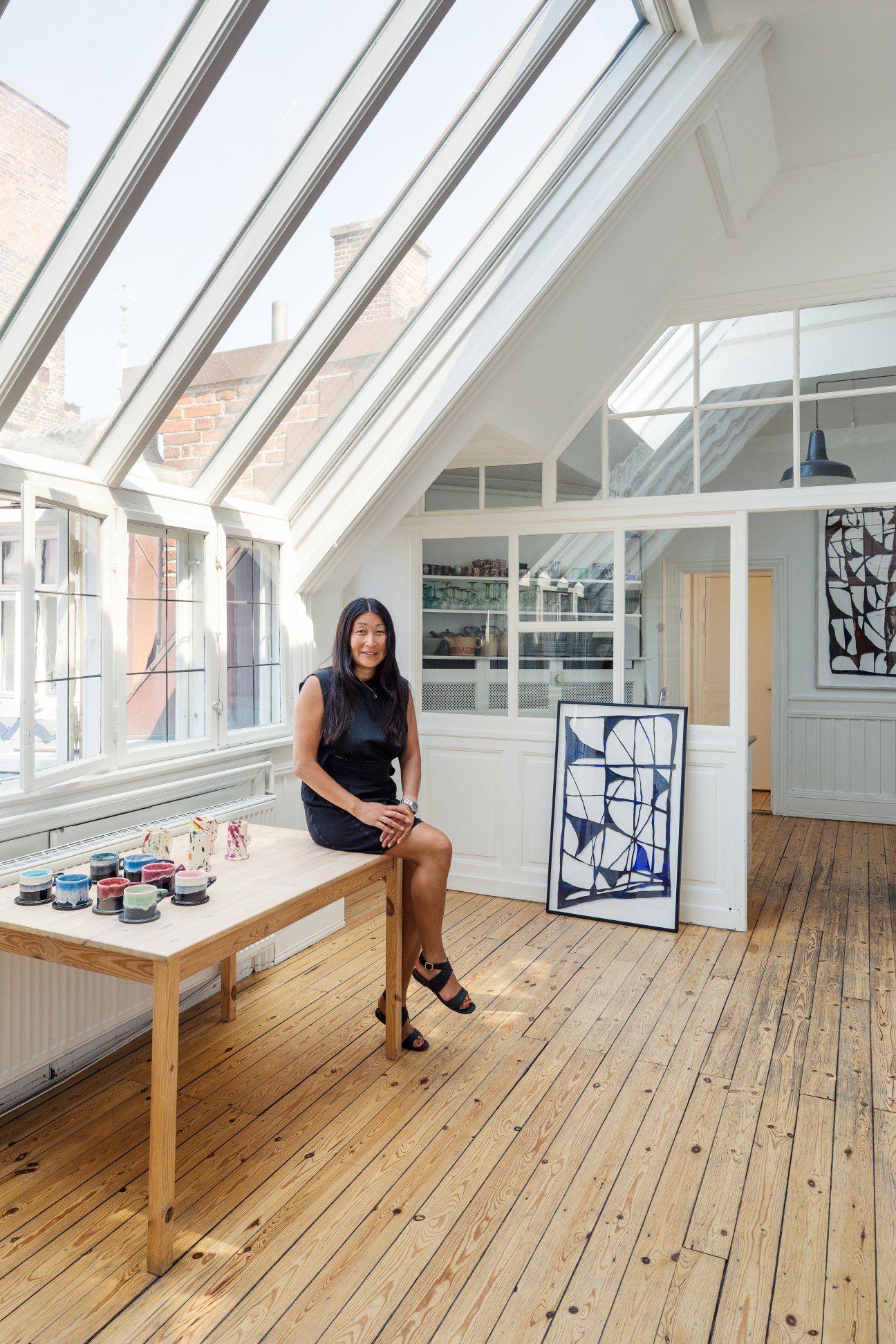 Anneli Häkkinen sits at a wooden table in her art gallery Les Gens Heureux, with skylights and brick walls visible above.