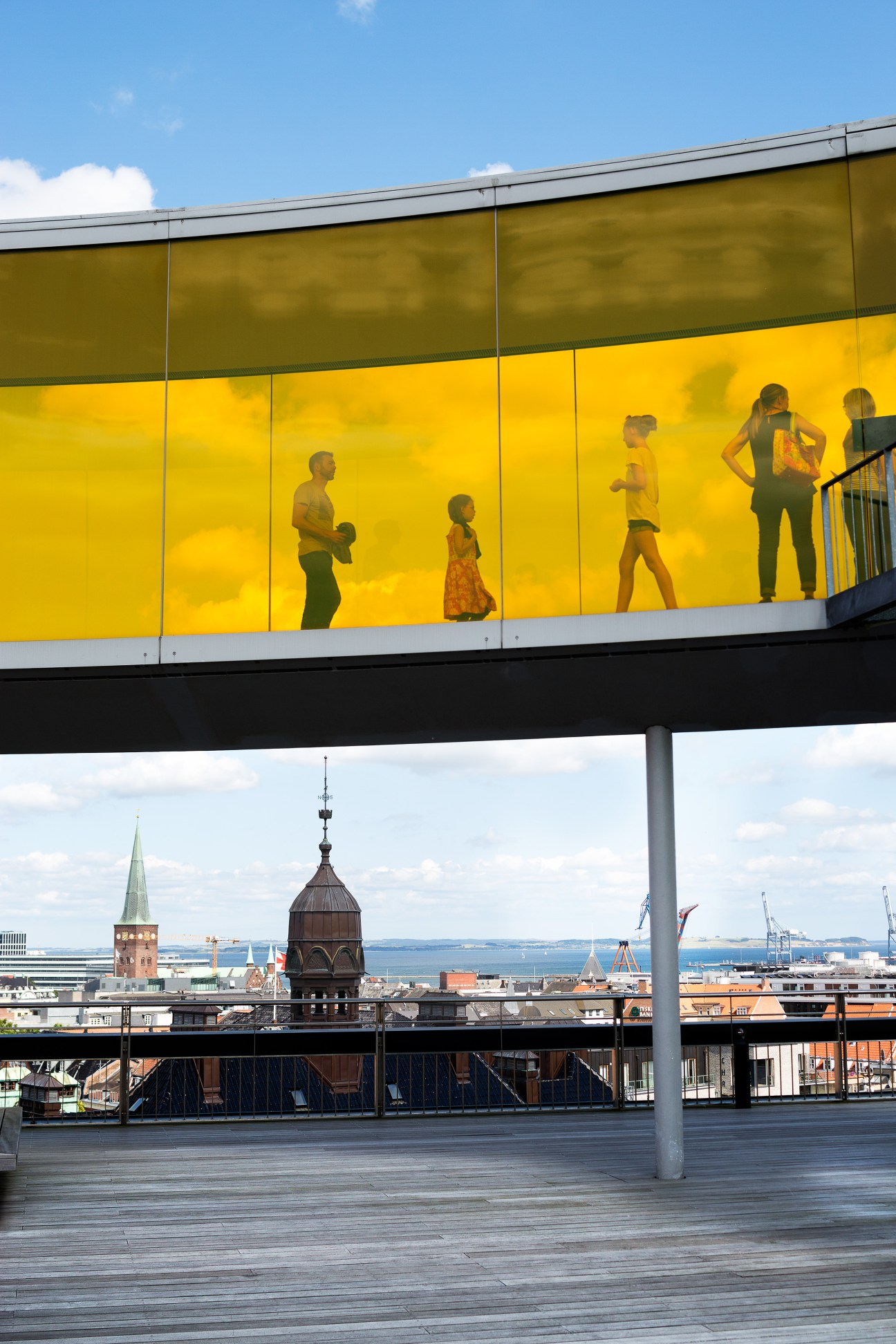 People silhouettes visible through yellow-tinted glass panels with city skyline and harbor visible below.