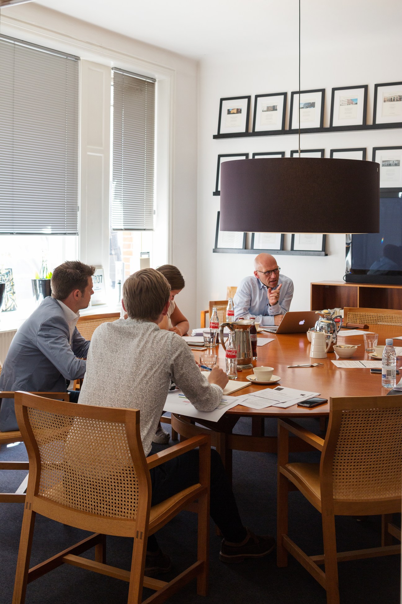 Four people in a business meeting around a wooden table in a modern office with framed artwork on the walls.