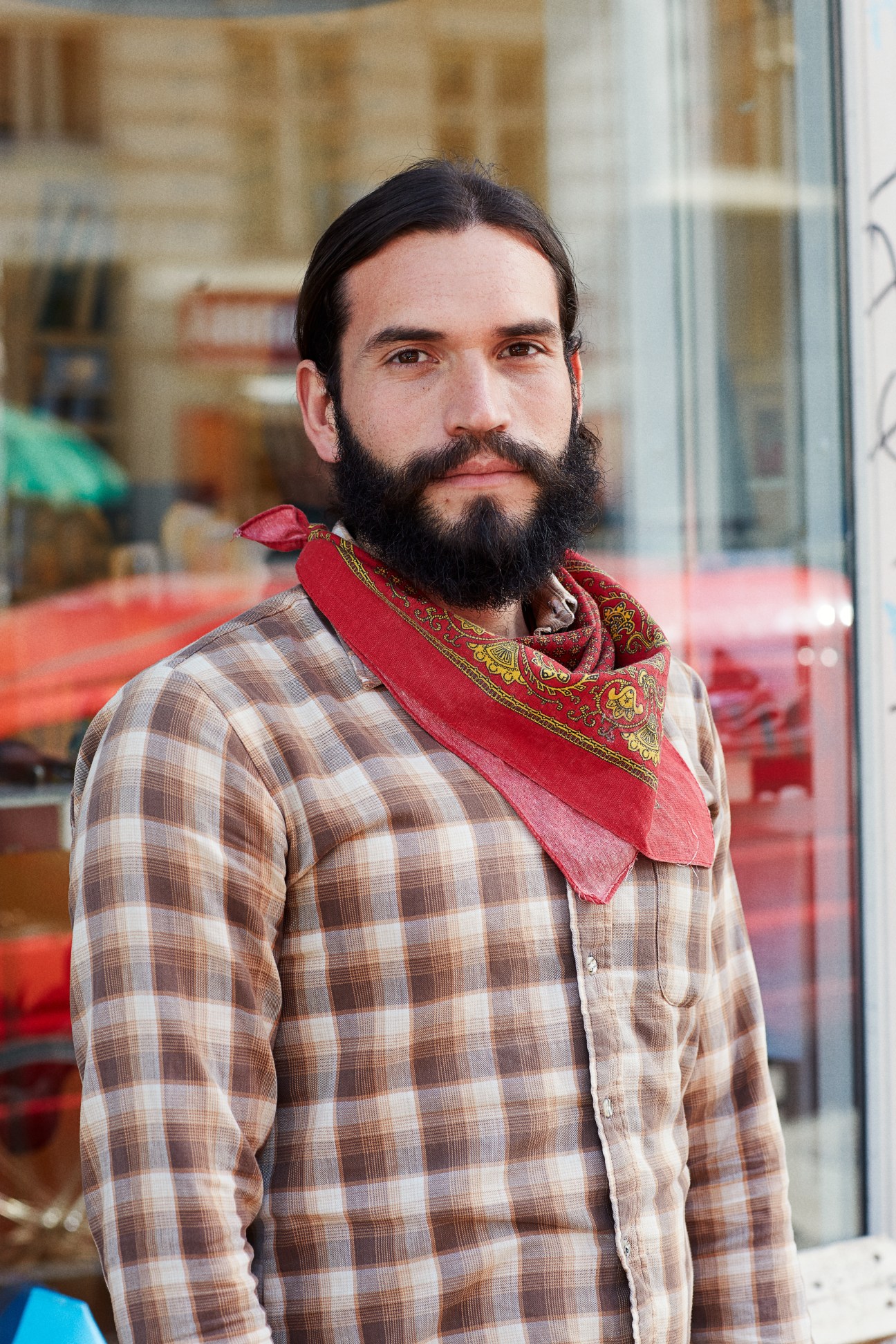 Bearded man in plaid shirt and red patterned scarf outside storefront.