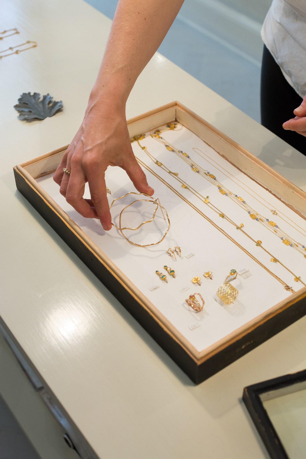 A hand reaches toward a display tray of gold jewelry pieces including rings, necklaces, and bracelets.