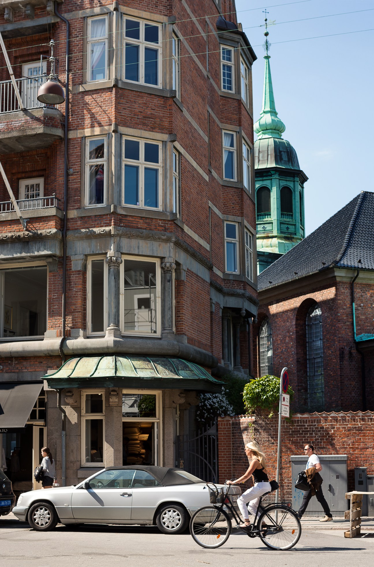 Red brick buildings with bay windows and a green-domed church tower in Copenhagen's Store Strandstraede, with pedestrians and a car on the street below.