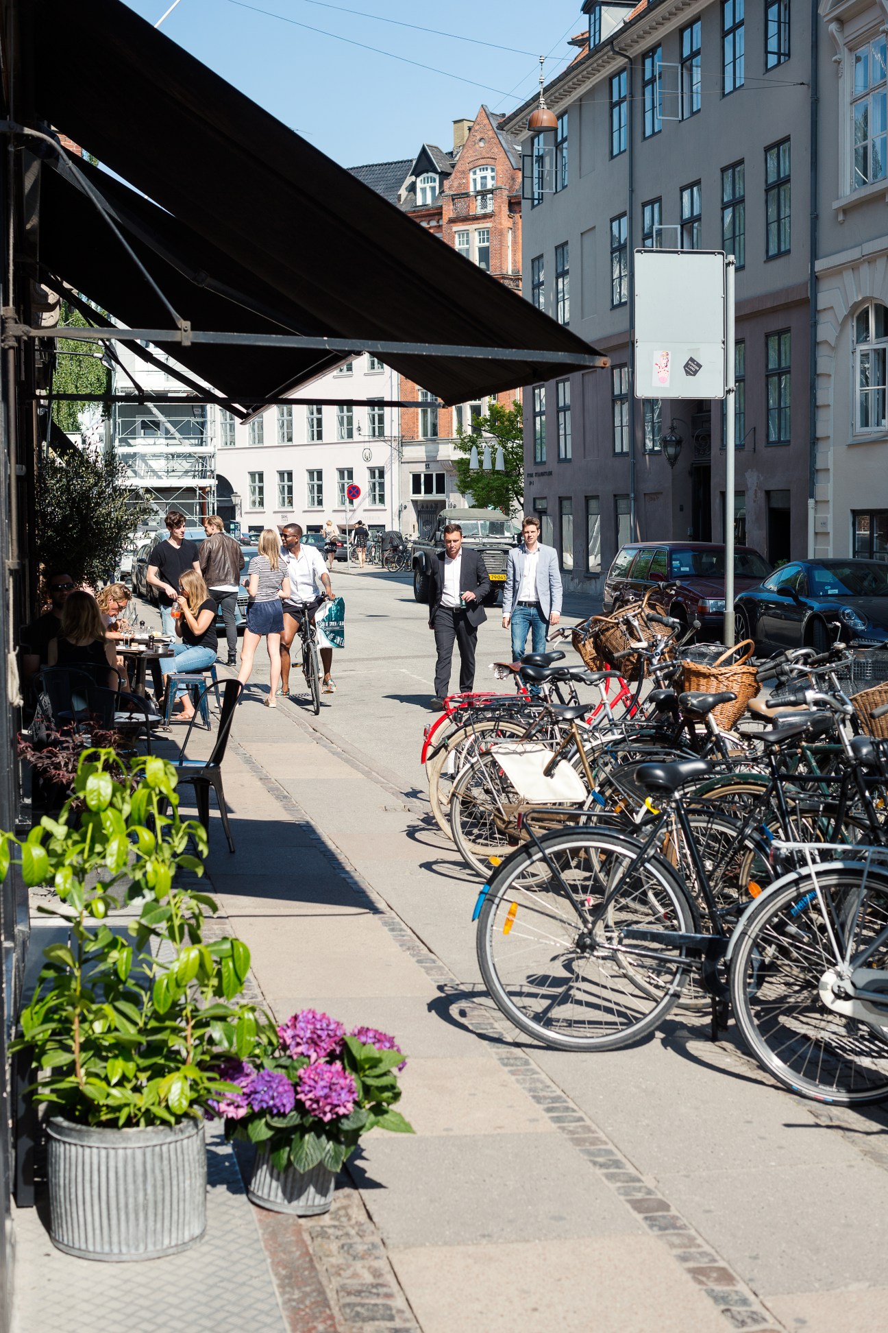 A Copenhagen street lined with historic buildings, outdoor café seating, bicycles parked along the street, and pedestrians walking on a sunny day.