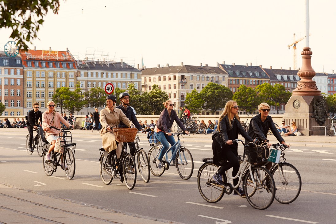 Multiple cyclists riding through a Copenhagen plaza with historic buildings and a clock tower in the background.