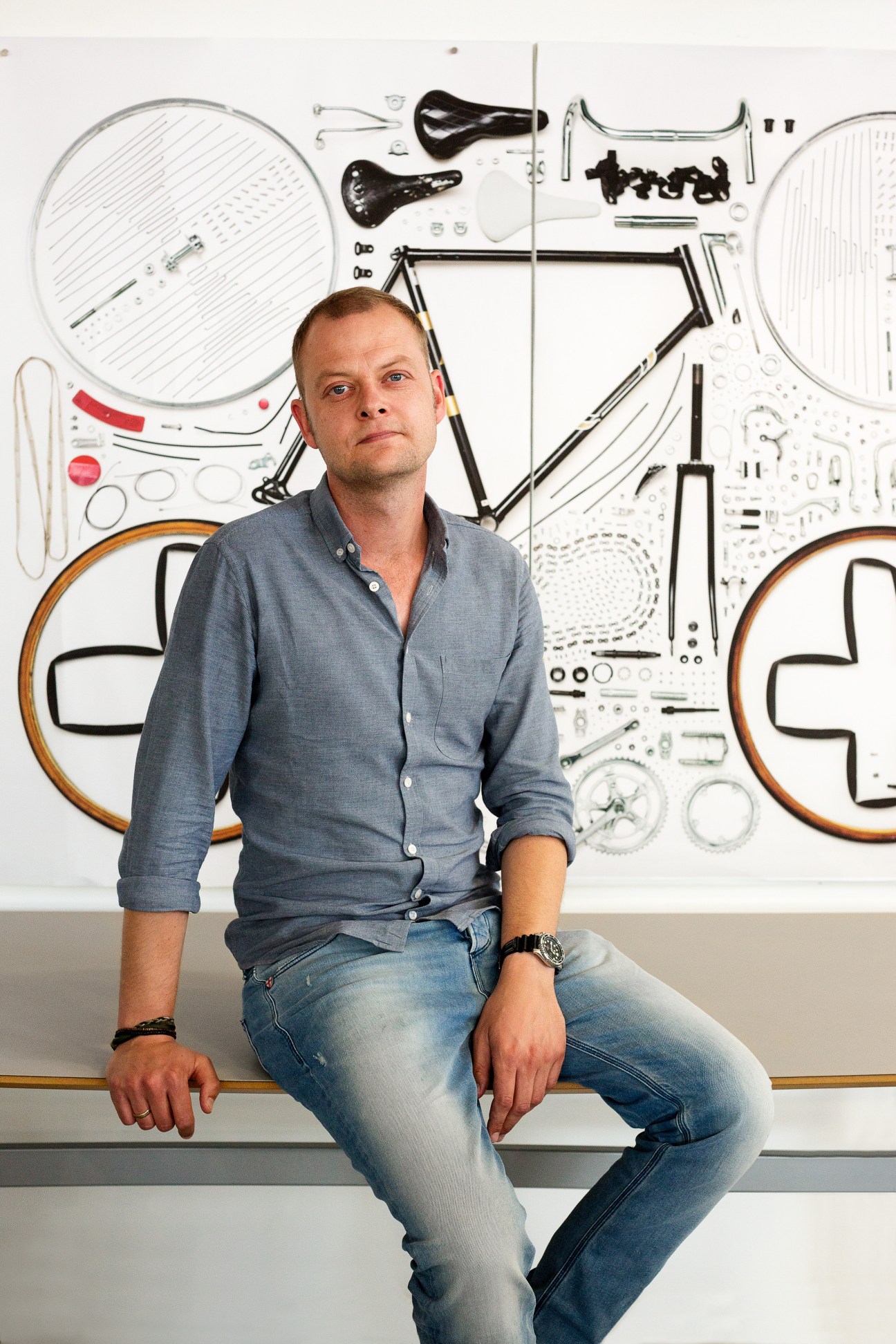 Man in blue shirt sitting in front of deconstructed bicycle parts wall display.