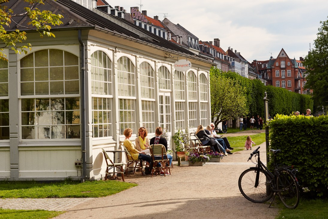 People sitting at outdoor café tables in a Copenhagen park with historic buildings and a bicycle nearby.