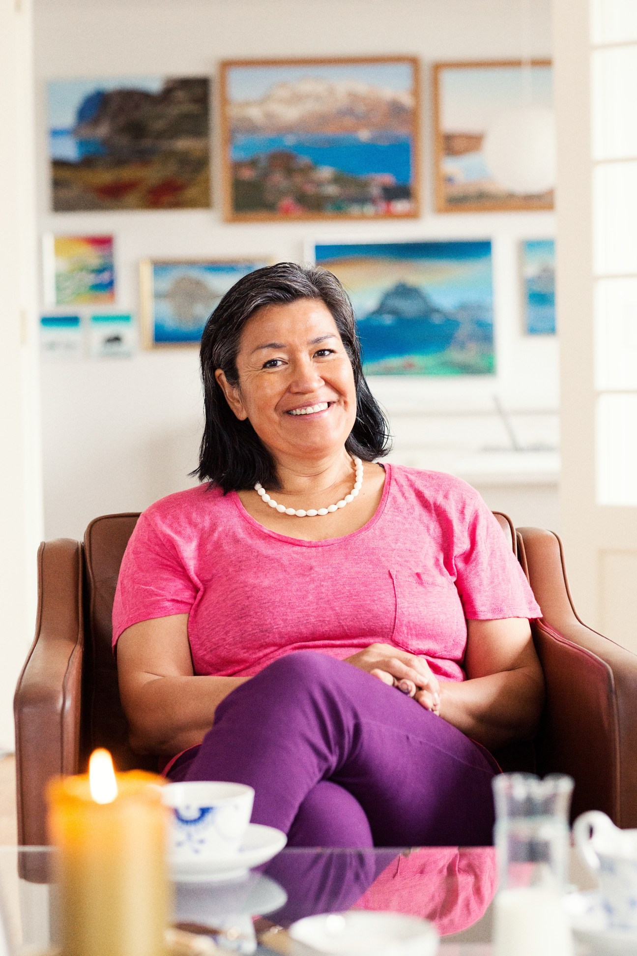 Aleqa Hammond, Greenland's prime minister, smiles while seated in a brown chair at her home in Nuuk, with framed landscape photographs on the wall behind her.