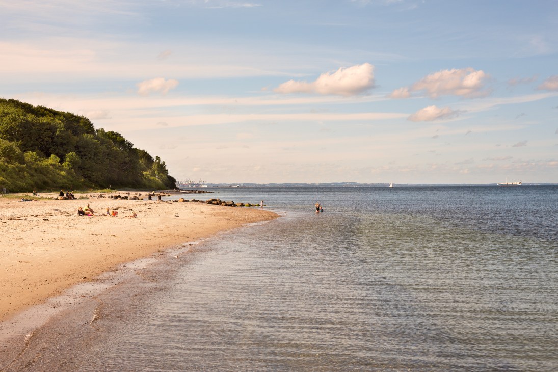 Sandy beach with visitors, forested cliffs and calm sea under cloudy sky.