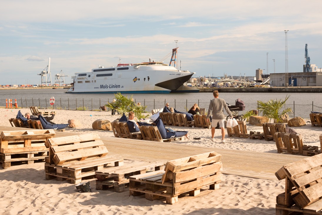 People relax on wooden pallet furniture at a sandy harbourside beach with a ferry ship and port cranes in the background.