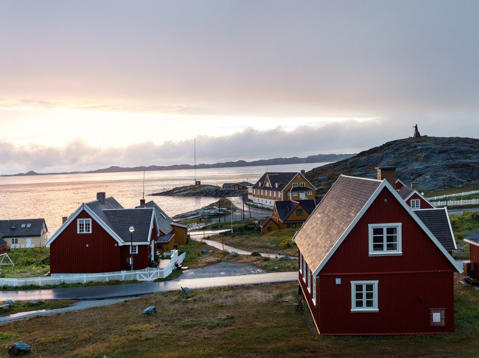 Red wooden houses with white-trimmed windows line a Greenlandic coastal settlement overlooking a fjord at dusk.