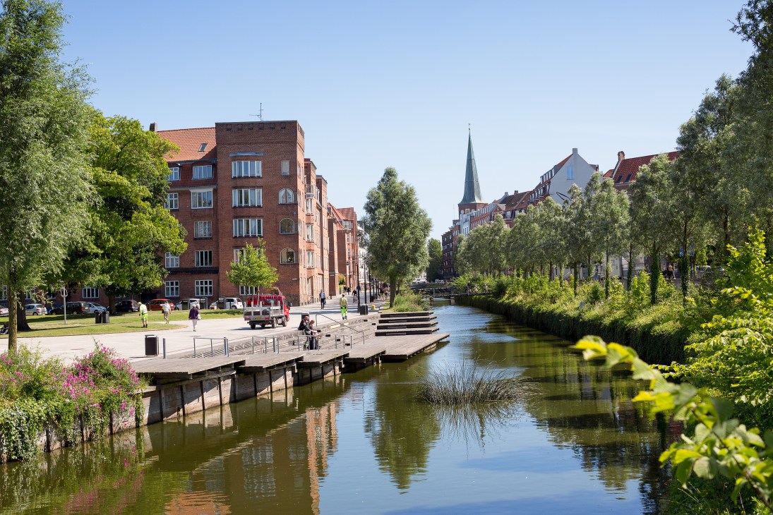 Tree-lined waterway with wooden boardwalk, red brick buildings, and church spire in Aarhus, Denmark