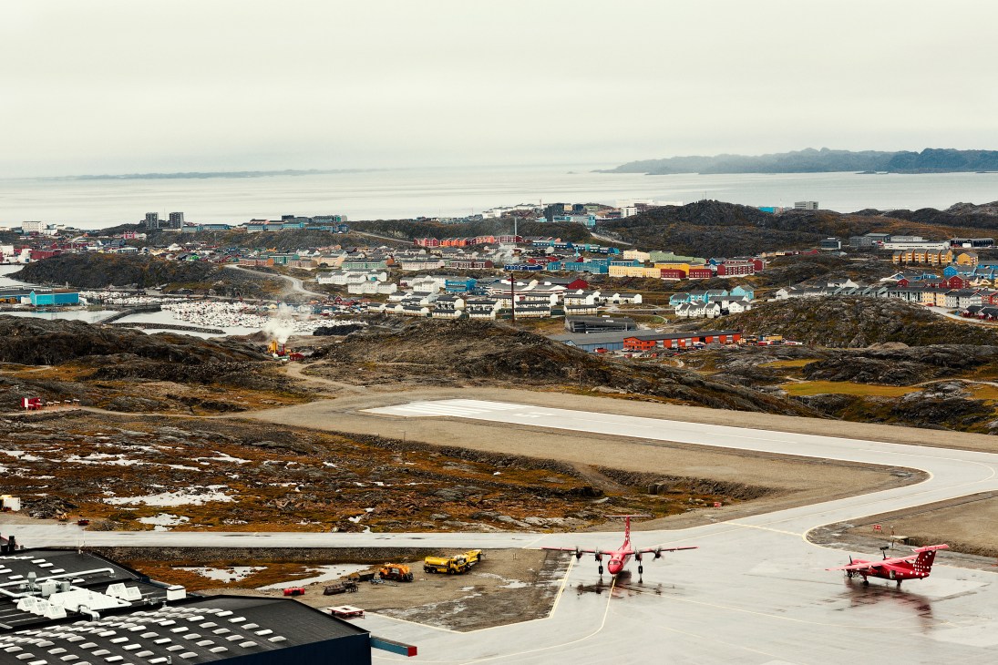 Aerial view of Nuuk airport with red aircraft on runway, colorful buildings and fjord in background.